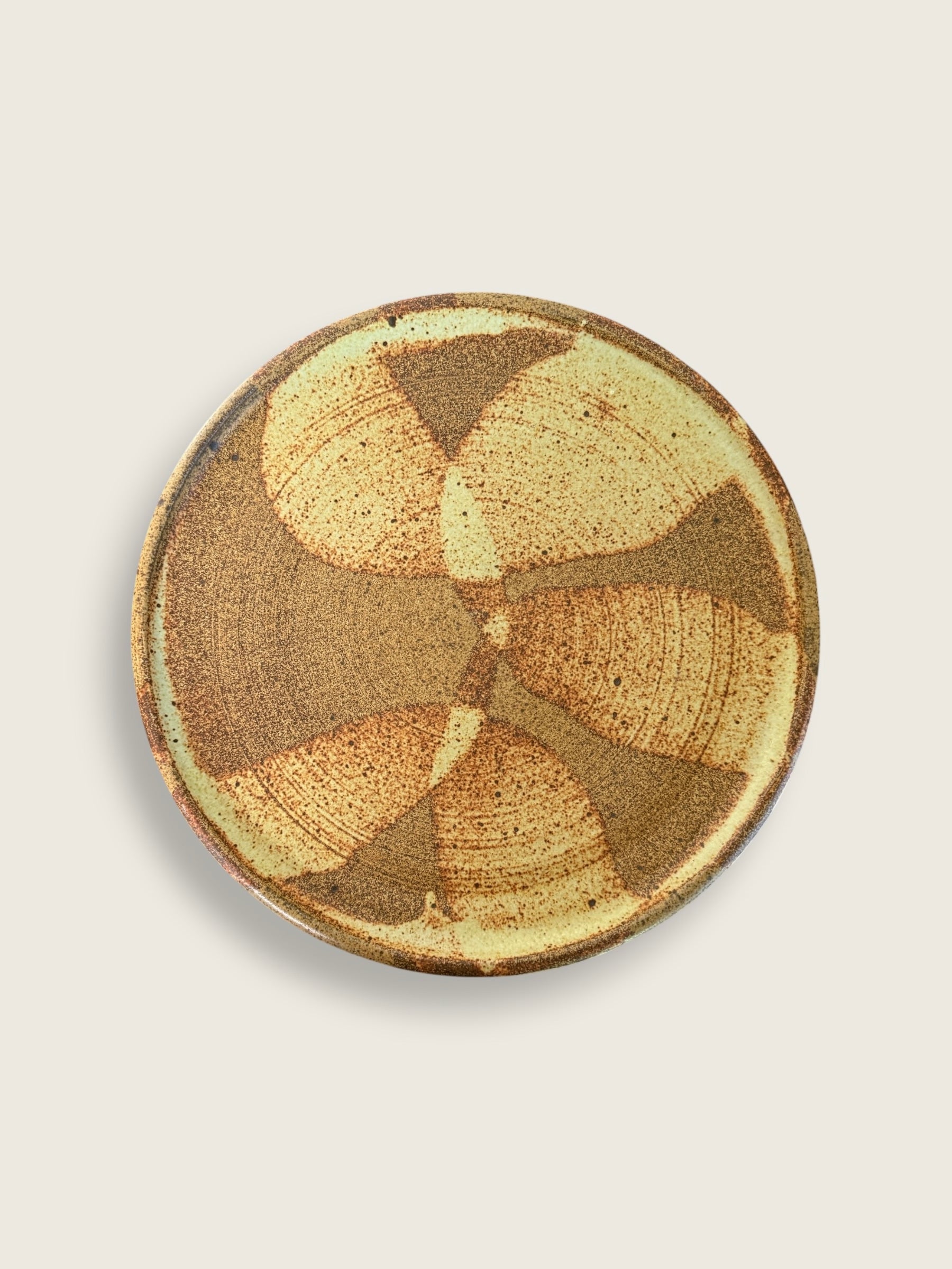 A round ceramic plate with a textured, hand-painted abstract pattern in earthy brown, tan, and cream tones, shown from a top-down view on a light beige background.