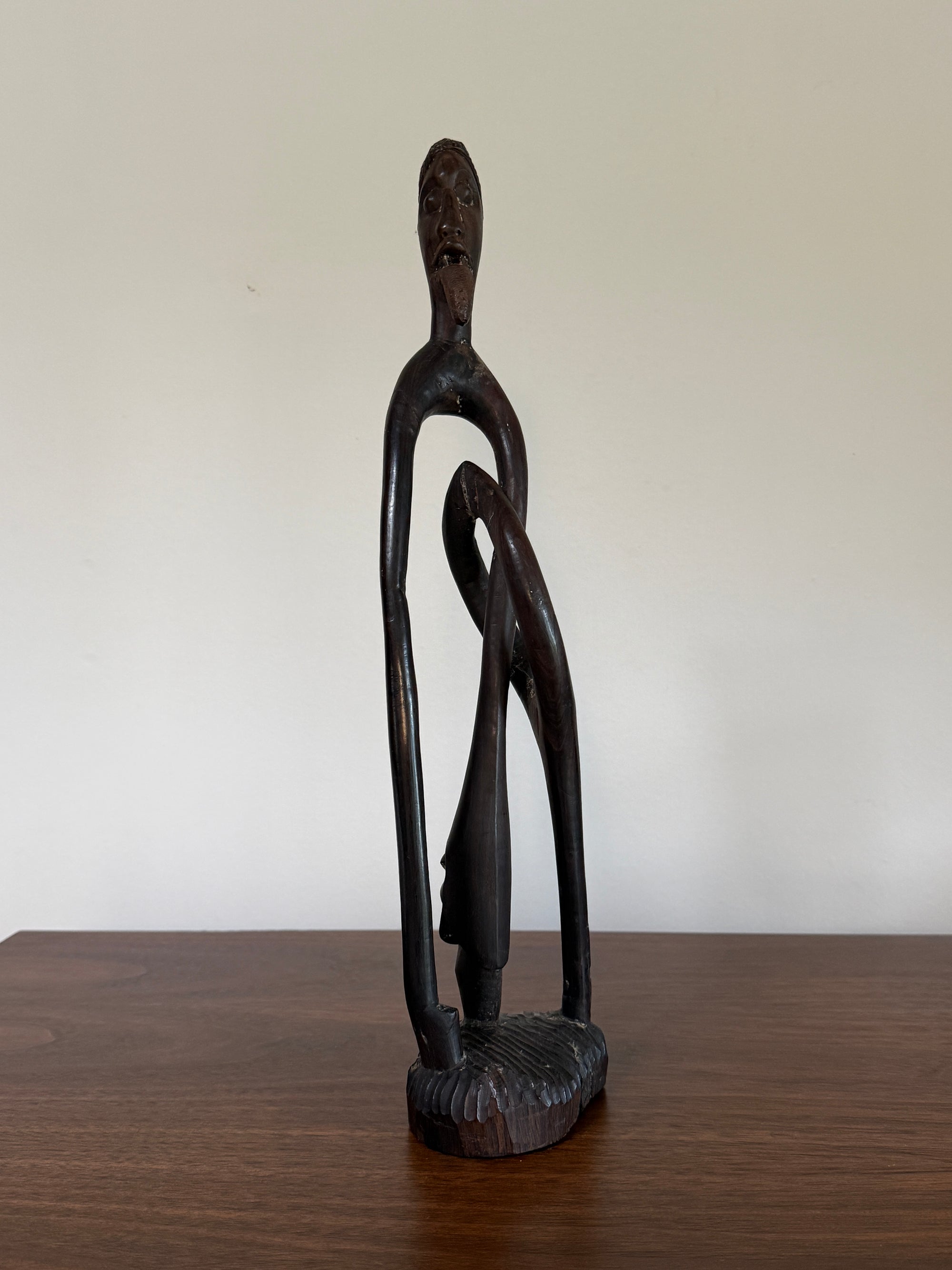 A dark carved wooden figurative sculpture of a stylized human figure standing on a base, placed on a wooden tabletop against a plain off-white wall.