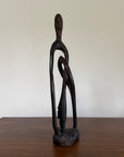 A dark carved wooden figurative sculpture of a stylized human figure standing on a base, placed on a wooden tabletop against a plain off-white wall.