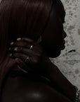 A close-up side profile of a woman with long dark brown hair, wearing silver rings on her fingers and a silver chain bracelet, set against a textured light gray background.