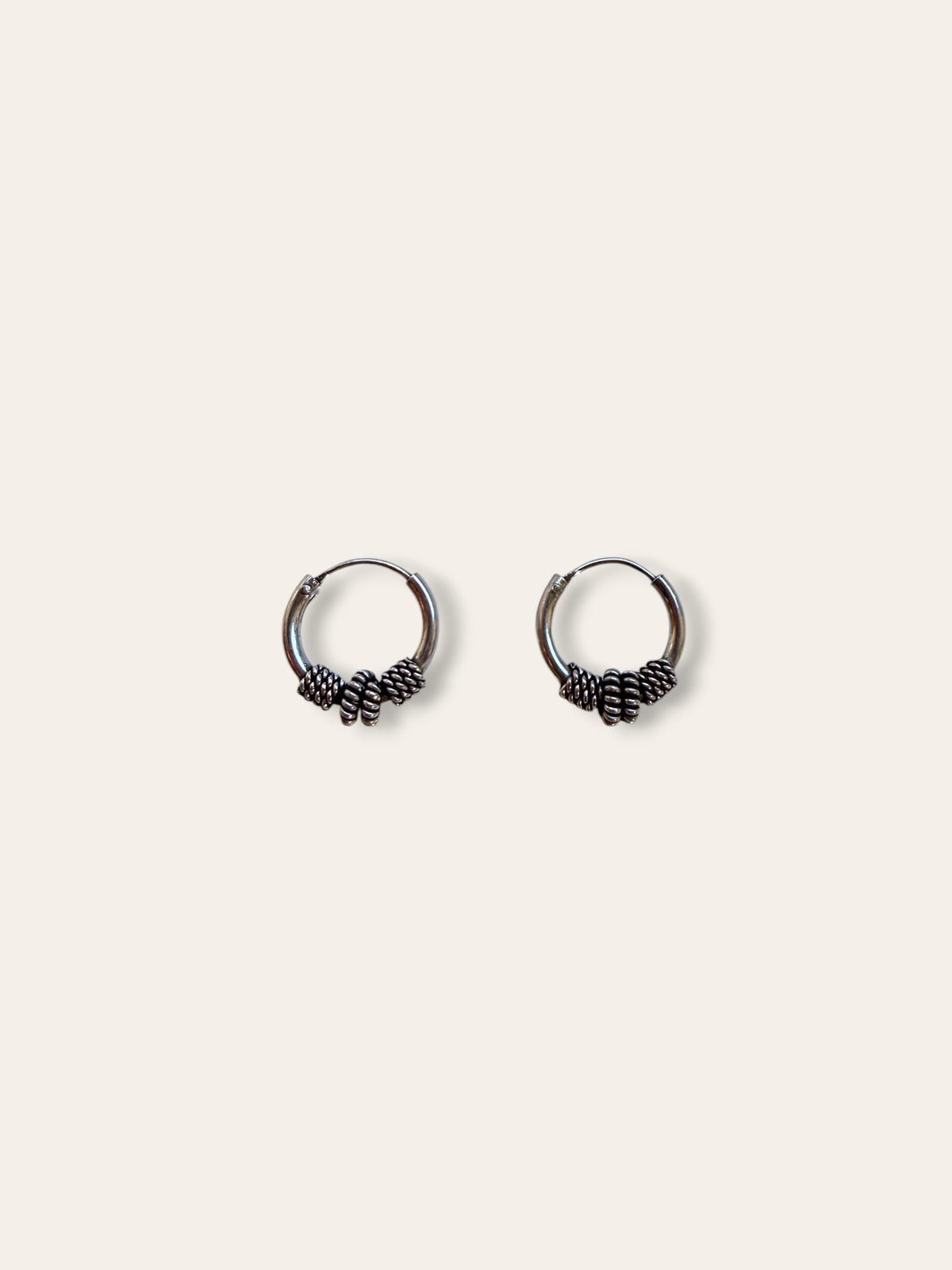 A pair of small silver hoop earrings with textured, knotted details at the bottom, displayed on a solid off-white background.