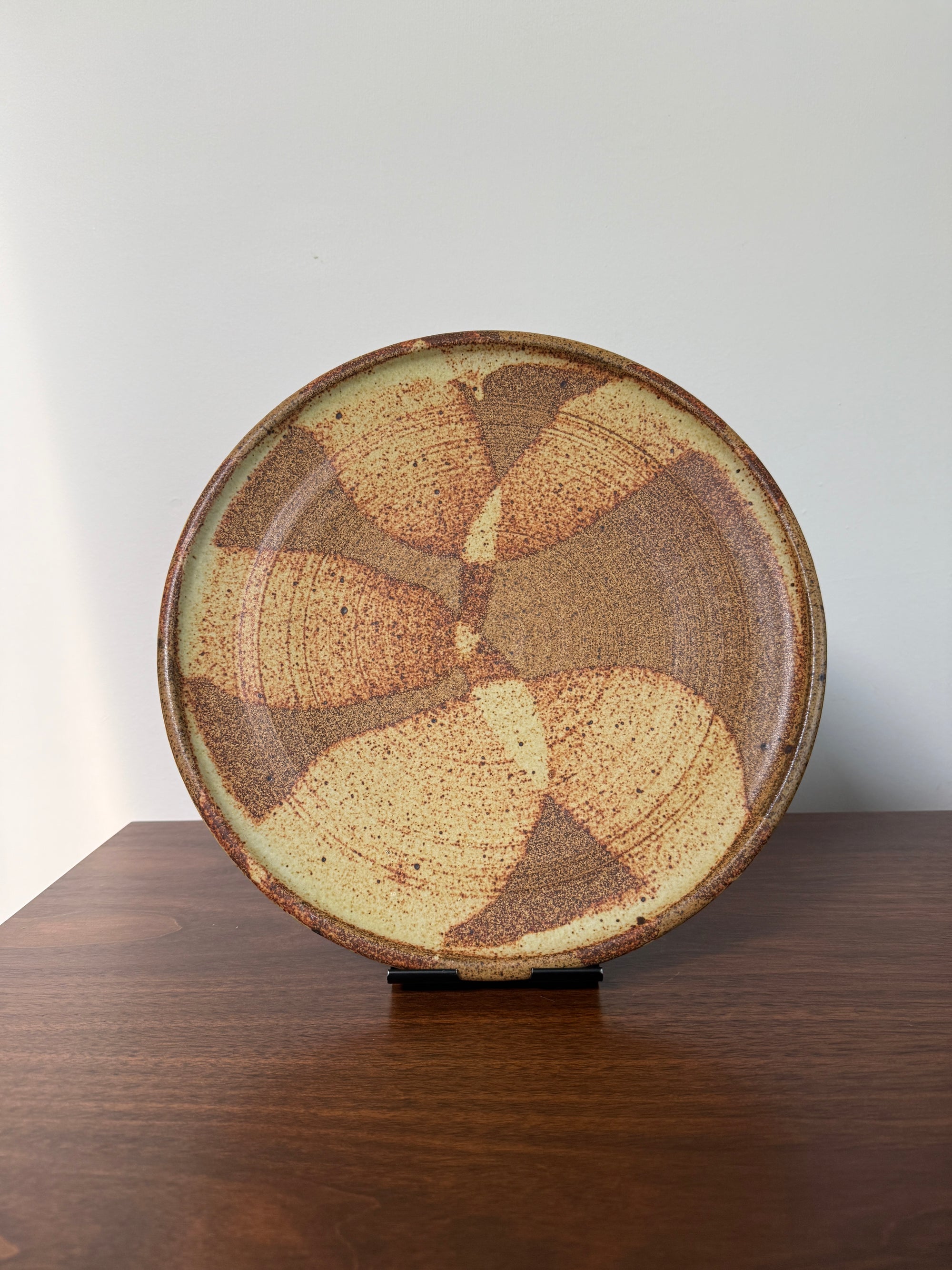 A round ceramic plate with a textured, hand-painted abstract pattern in earthy brown, beige, and tan tones, displayed on a dark wooden surface against a plain light gray wall.