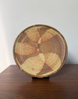 A round ceramic plate with a textured, hand-painted abstract pattern in earthy brown, beige, and tan tones, displayed on a dark wooden surface against a plain light gray wall.
